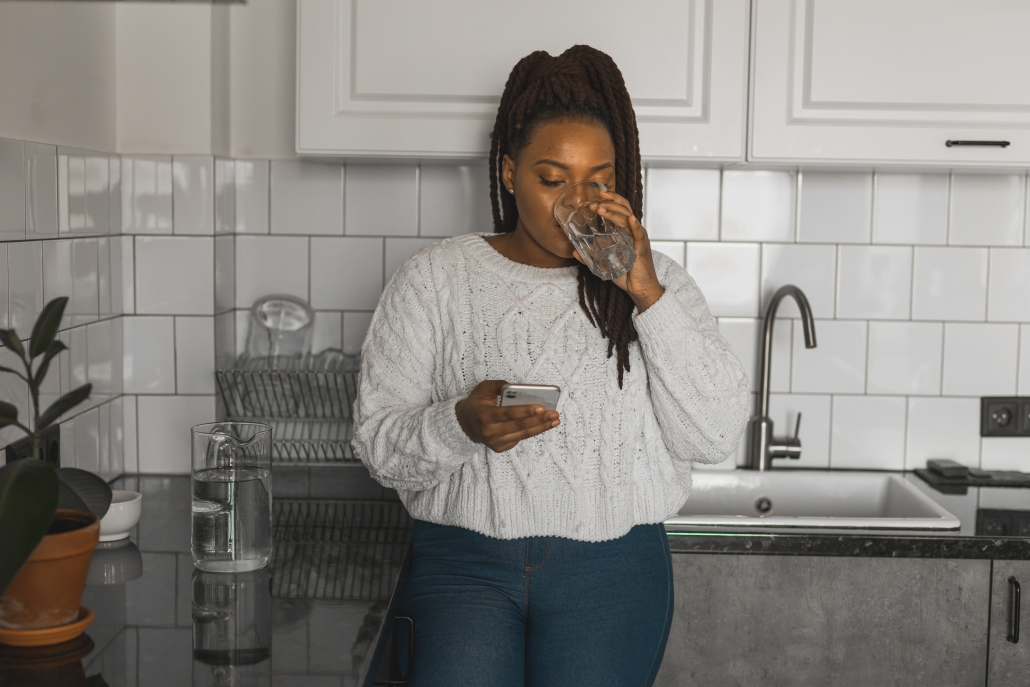 woman drinking a glass of water in her kitchen while looking at phone